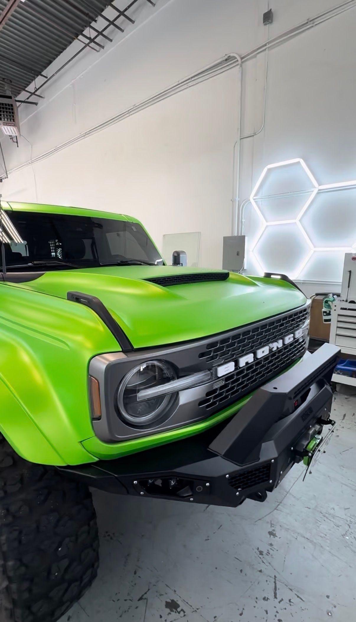 Bright green off-road truck inside a garage, viewed from the front-left with neon hexagon wall lights.