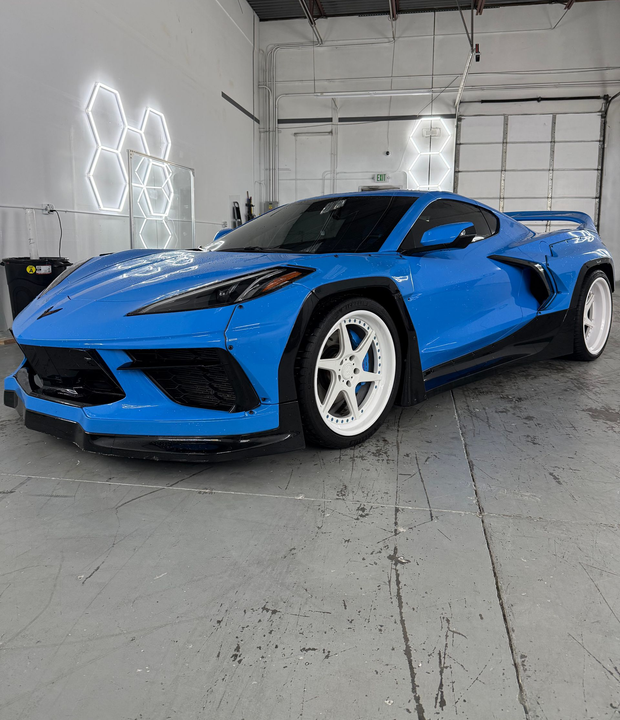 Blue Chevrolet Corvette in a garage, angled front-left view with white wheels and LED wall lights.