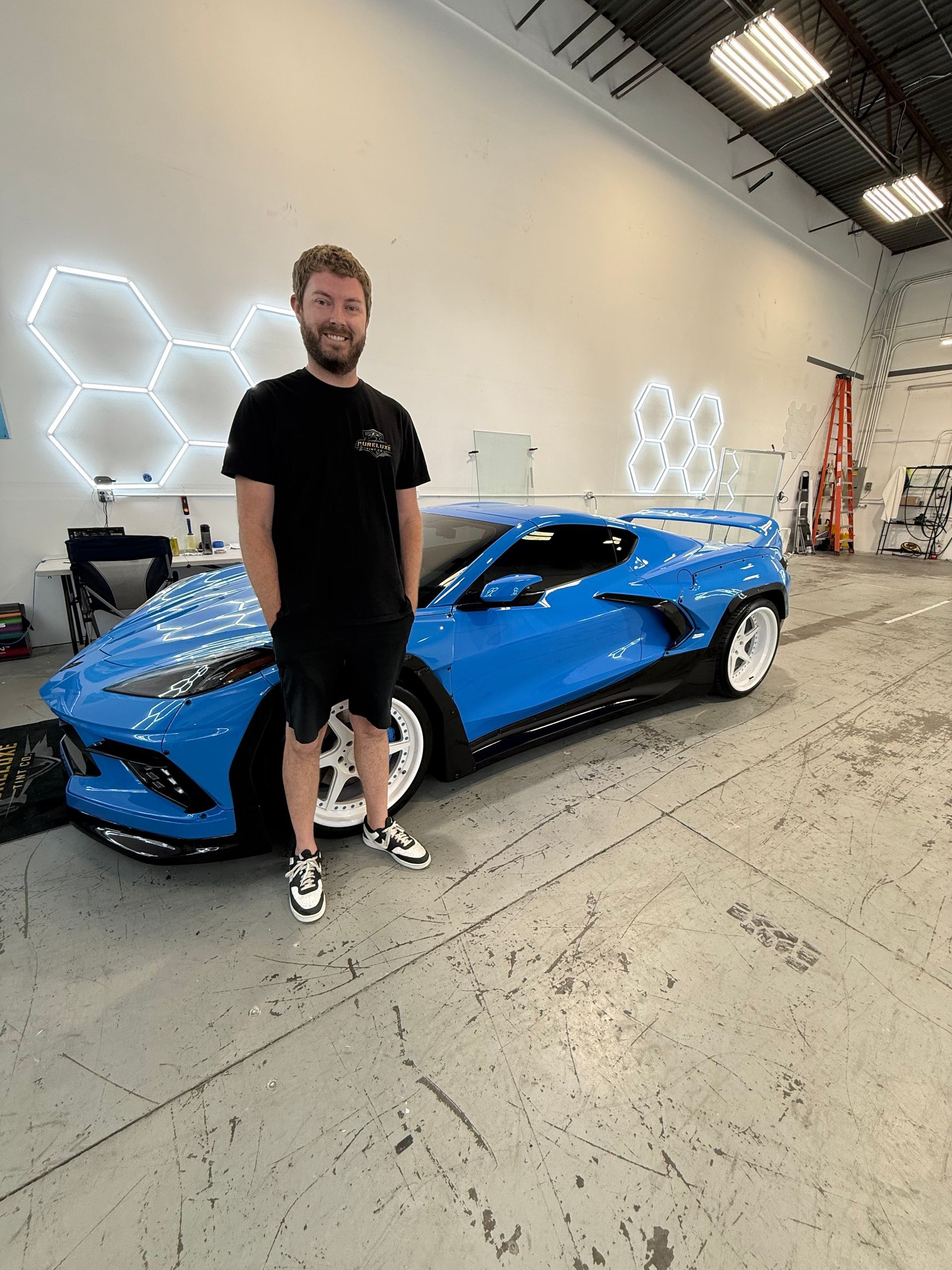 Man standing beside a blue sports car in a bright garage with hexagon wall lights
