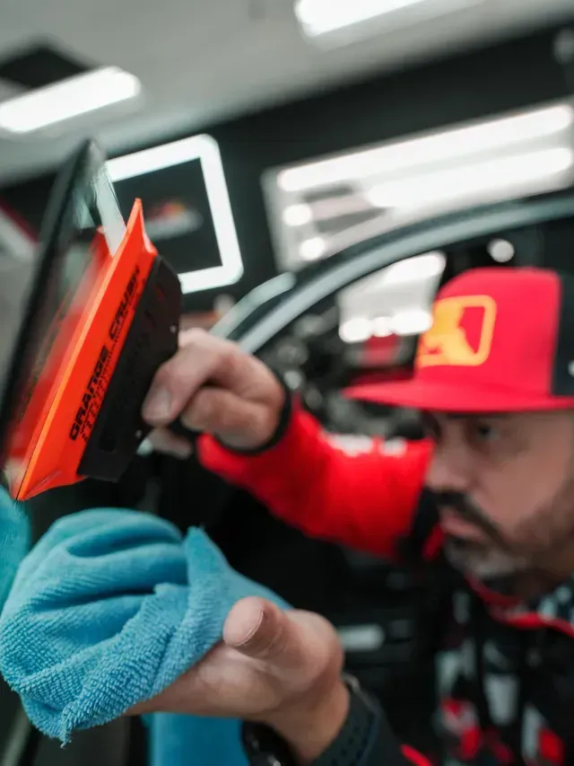 Technician using a blue cloth to clean an orange car window inside a garage