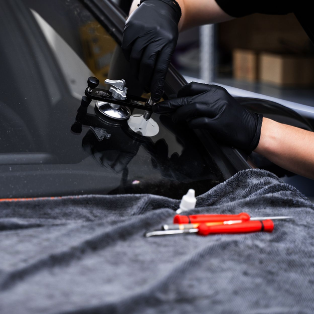 Gloved hands working on a car windshield with red tools on a gray cloth surface