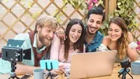 Four people laugh while looking at a laptop and small cameras, outdoors.