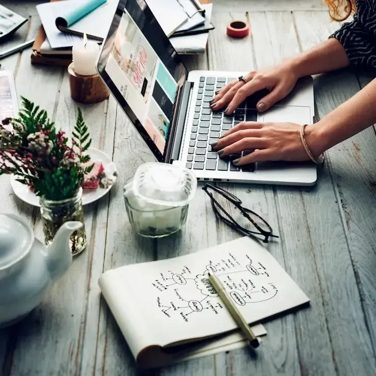 Hands typing on a laptop at a rustic desk with notebook, pen, coffee, and small plant.