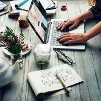 Hands typing on a laptop at a rustic desk with notebook, pen, coffee, and small plant.