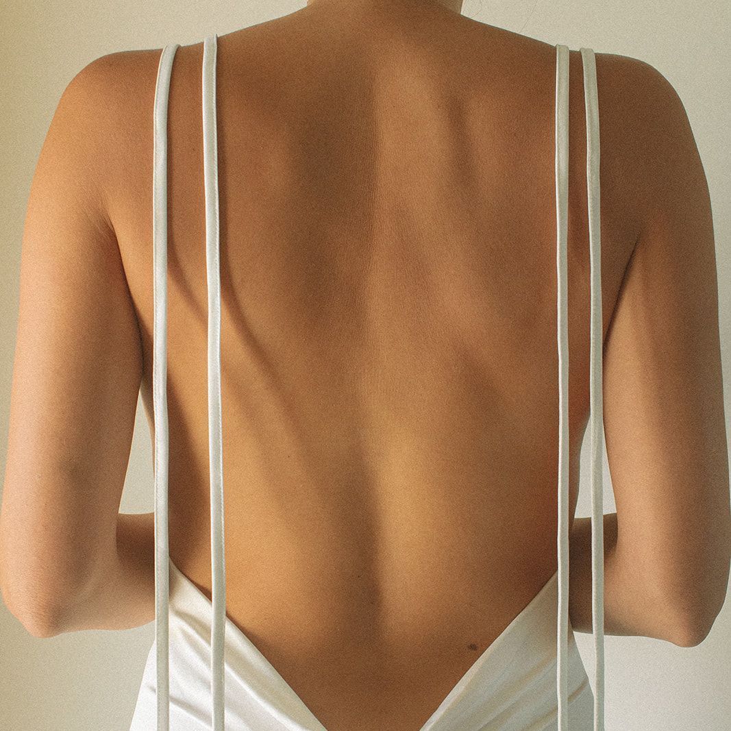 Back view of a person wearing a white dress with thin straps. Pale skin, neutral background.