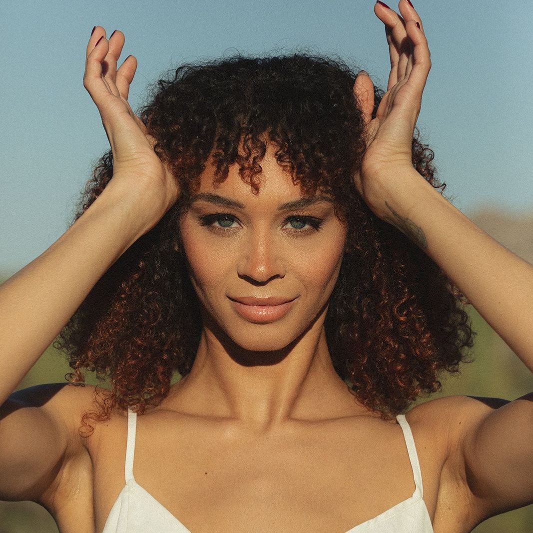 A woman with curly hair is wearing a white tank top