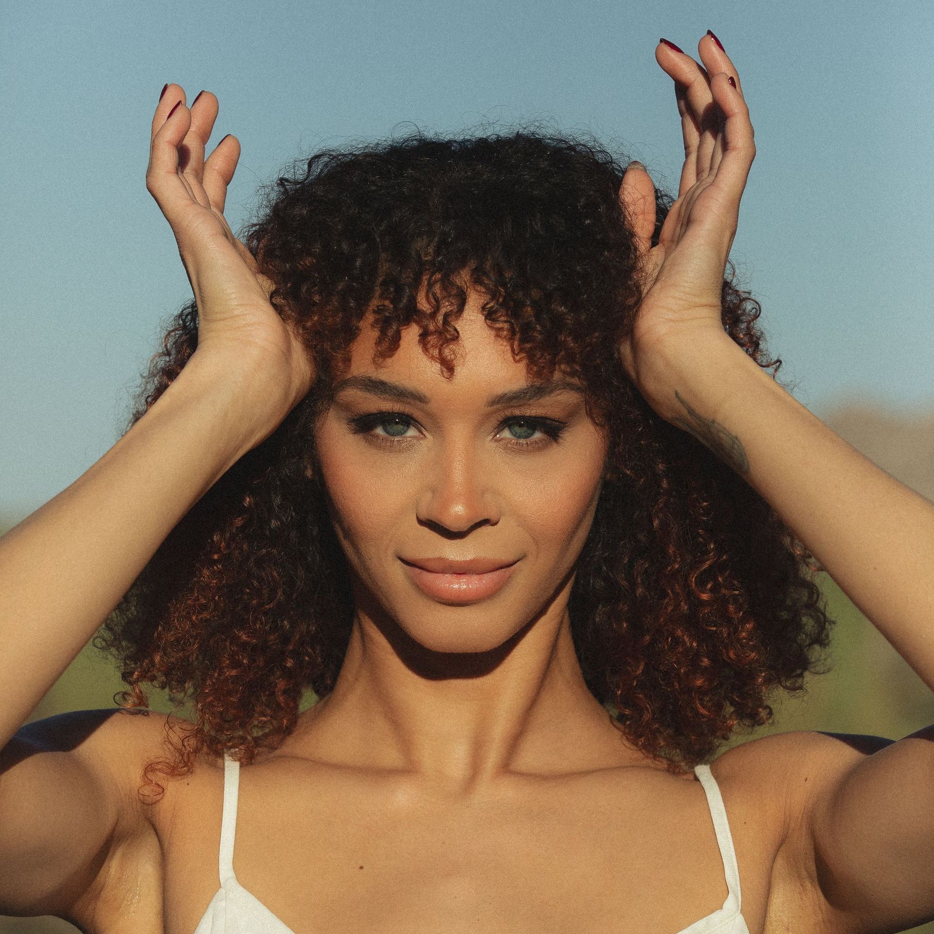 A woman with curly hair is wearing a white bra and holding her hands to her head.