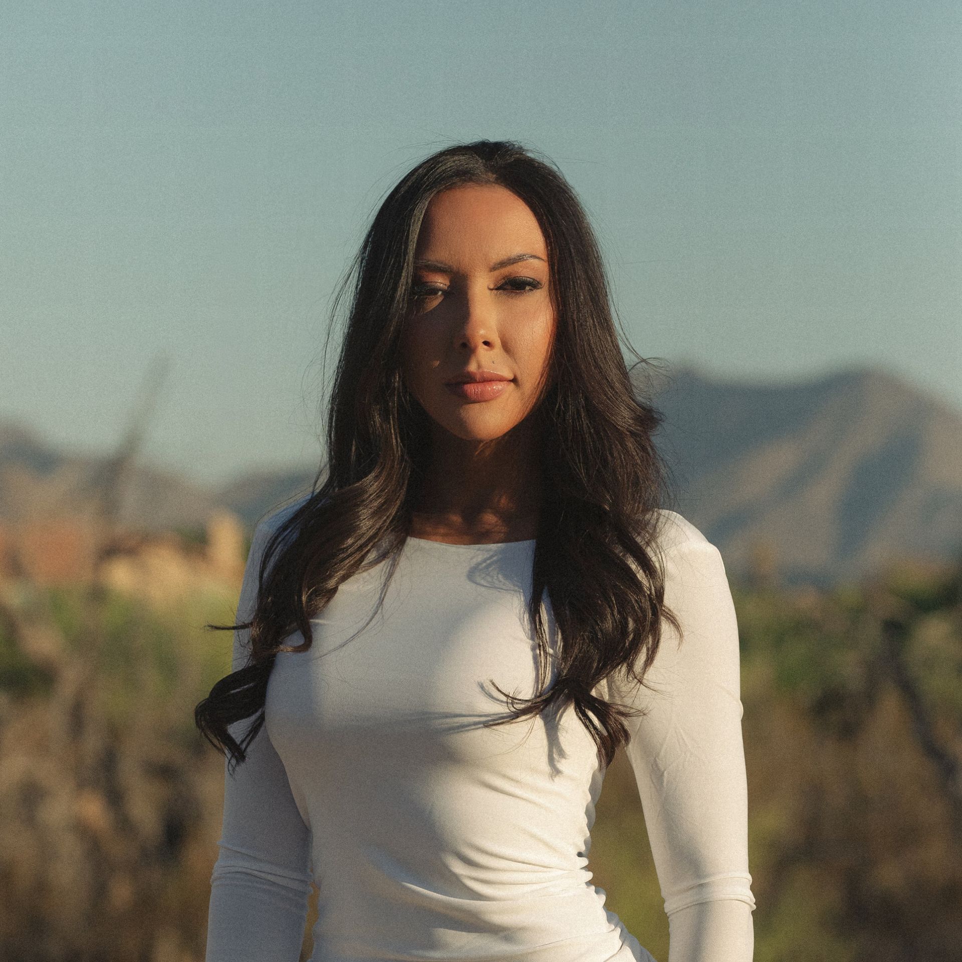 A woman in a white shirt is standing in a field with mountains in the background