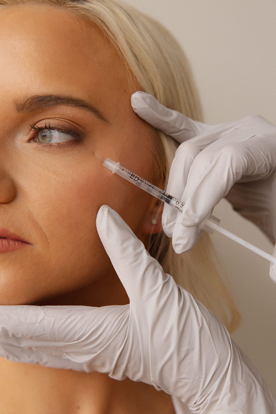 Gloved hands administering facial cosmetic injections to a blonde woman’s cheek