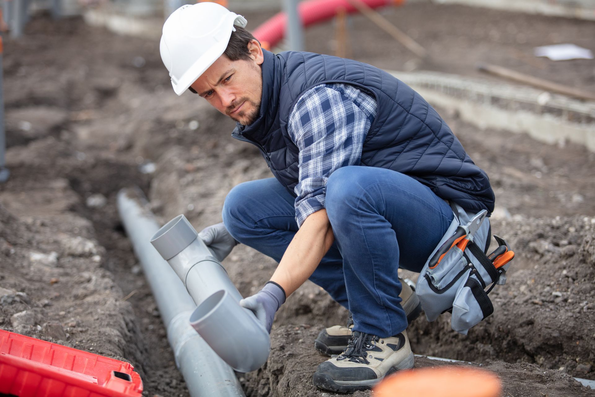 Worker in hard hat crouches by gray pipes at a construction site, using tools on the ground.