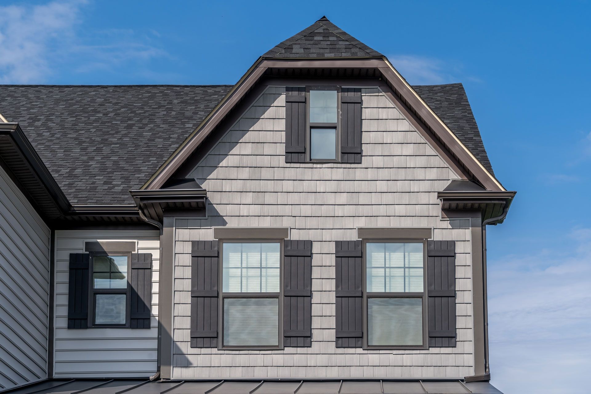 Close-up of a house with gray shingles, dark shutters, and a blue sky background.