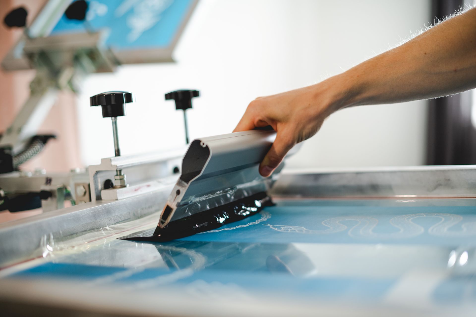 Hand pulling a squeegee across blue screen-printing ink on a print press