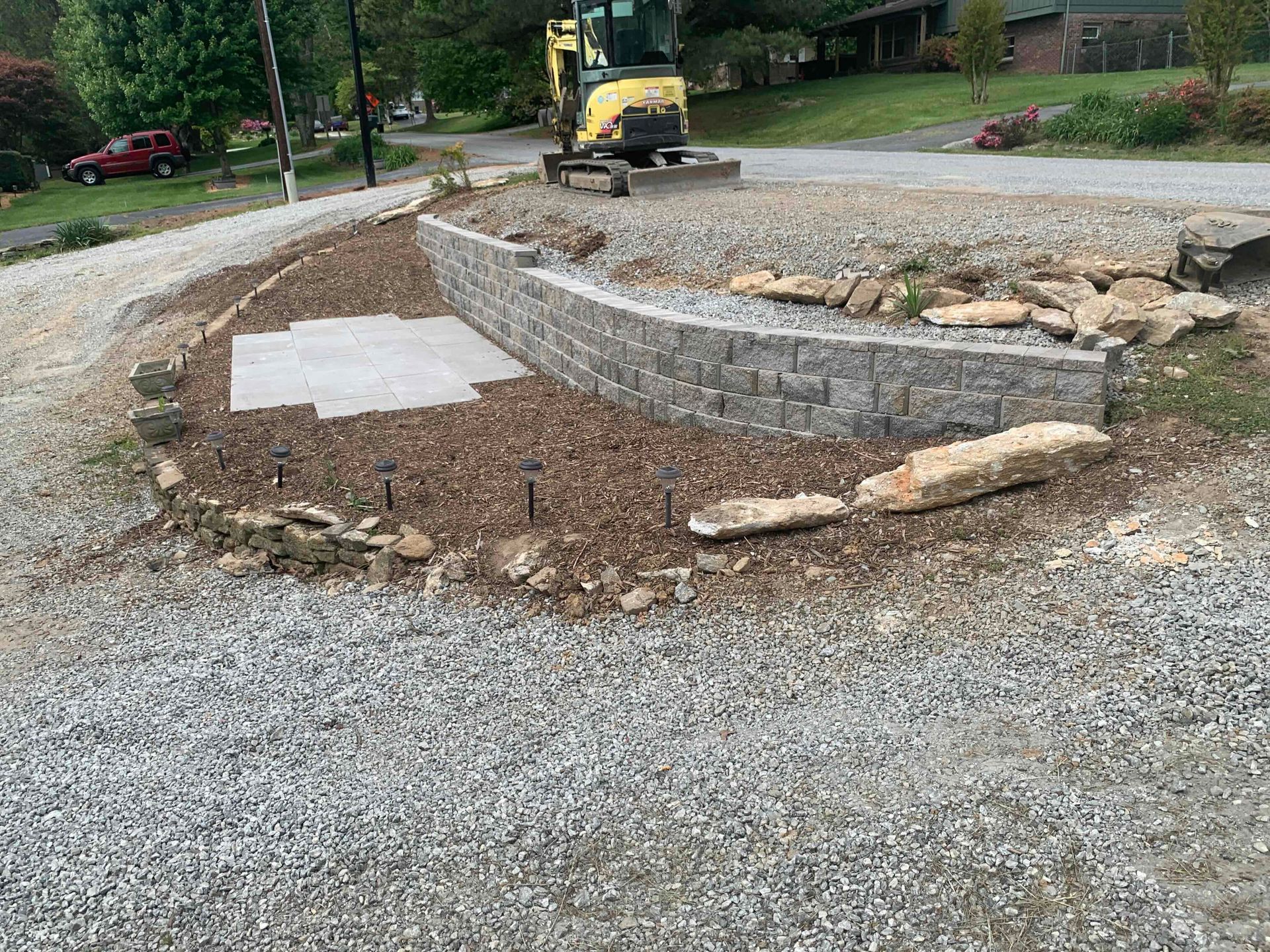 Curved stone retaining wall under construction beside a gravel driveway and a yellow skid steer.