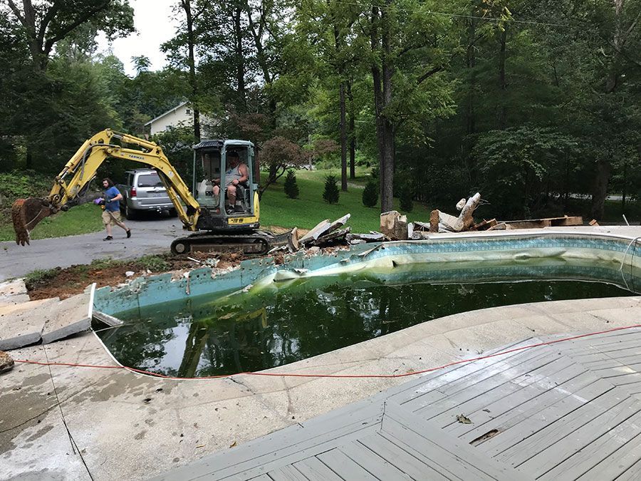 Backyard pool demolition with excavator, broken concrete, and drained water in wooded yard