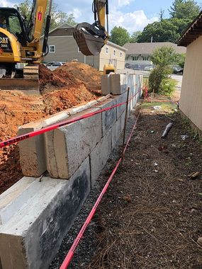 Excavator beside a new cinder block retaining wall at a muddy construction site, marked with red tape