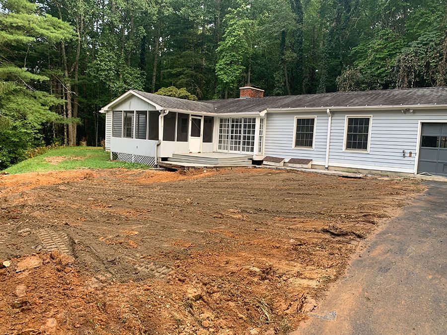 White single-story house with a screened porch and freshly cleared dirt yard, backed by trees
