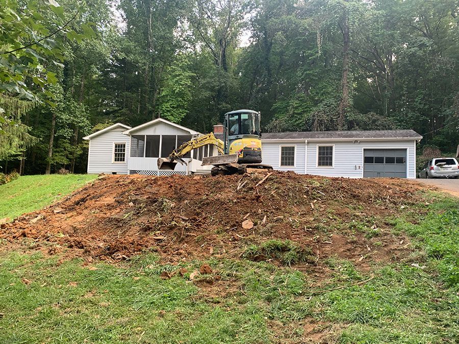 Bulldozer working on a dirt mound in front of a white house and garage in a wooded yard