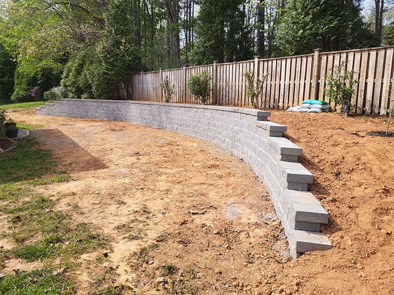 Curved gray retaining wall under construction in a wooded backyard with dirt and fence background