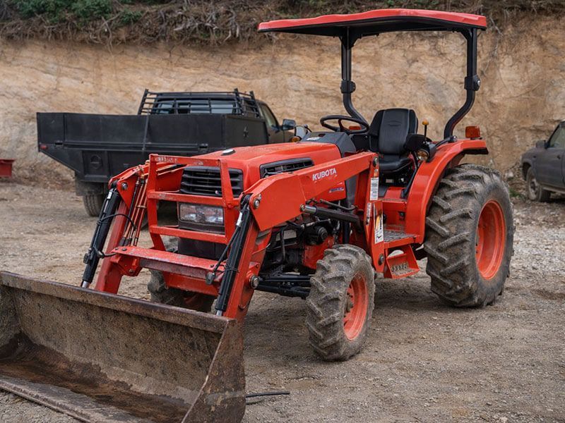 Red tractor with front loader and plow attachment on a dirt lot, beside a trailer and hillside.