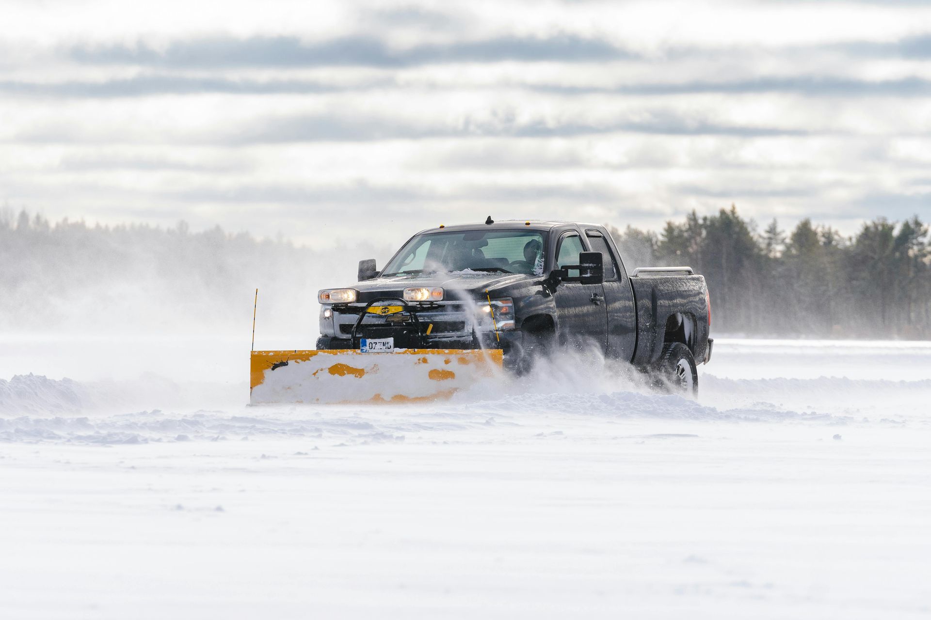 Black pickup plowing snow across a snowy field under a cloudy sky