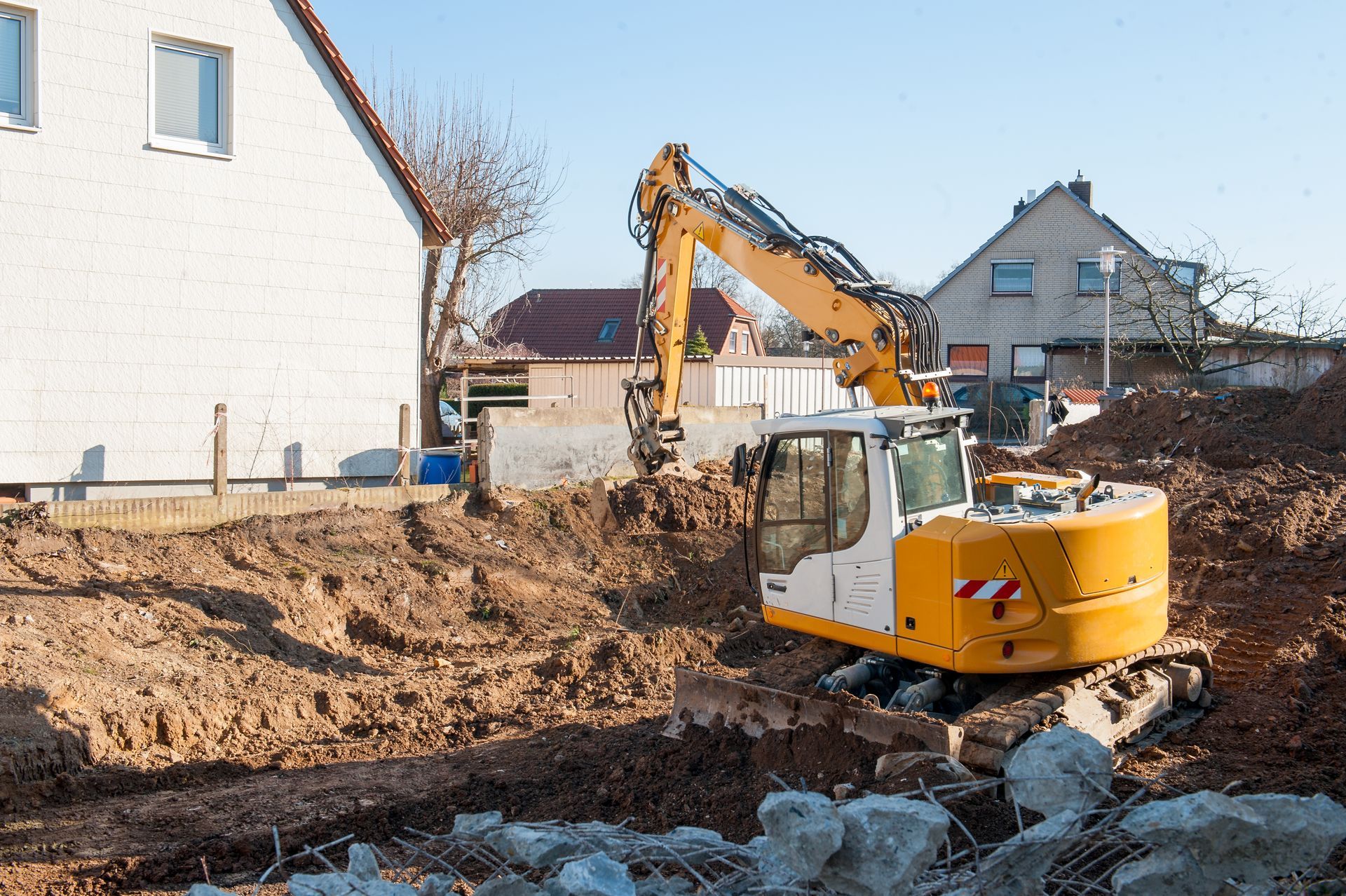 Excavator digging dirt at a house construction site beside a white building.