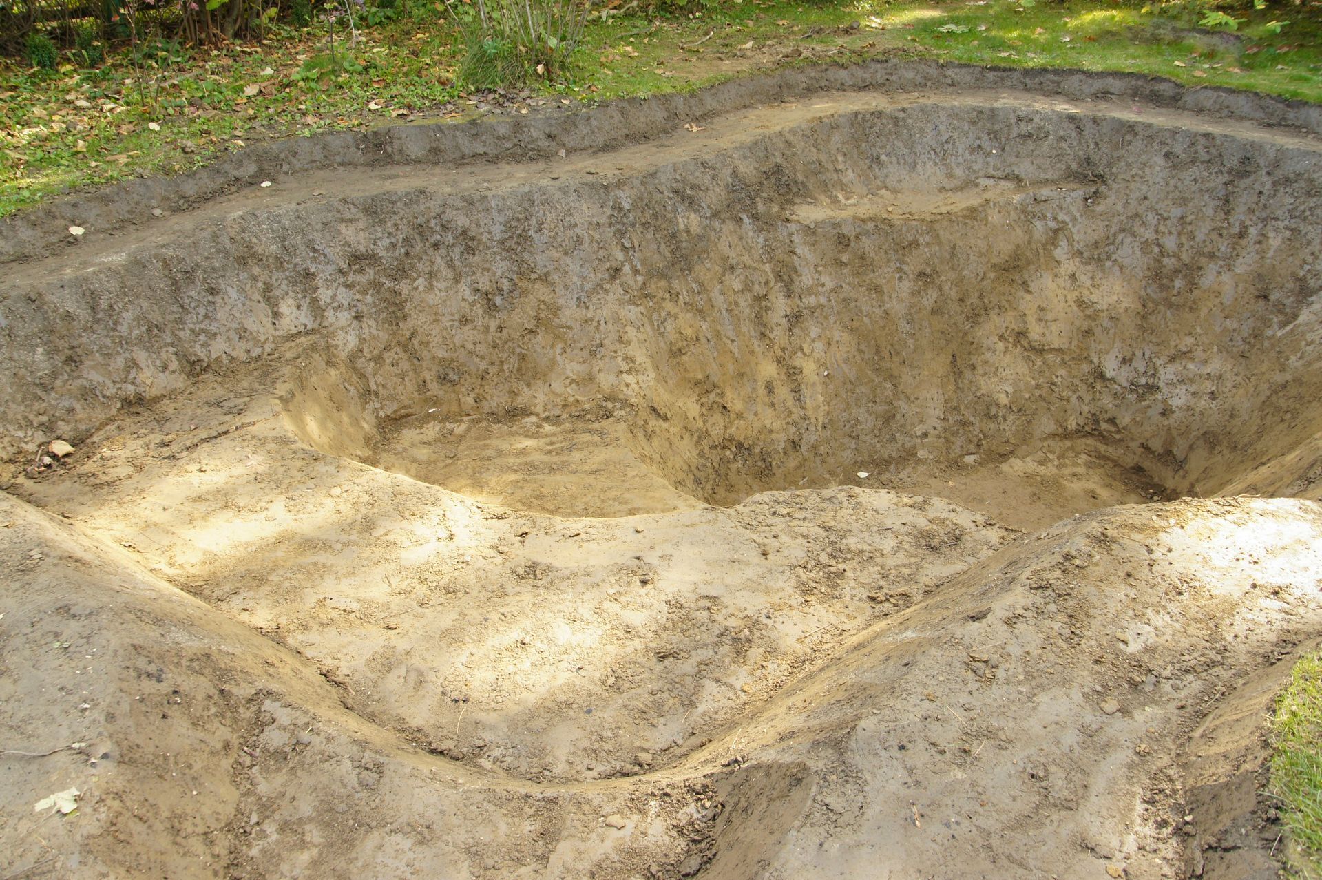 Large shallow dirt pit with sloped sides and pale sand, surrounded by grass and trees.