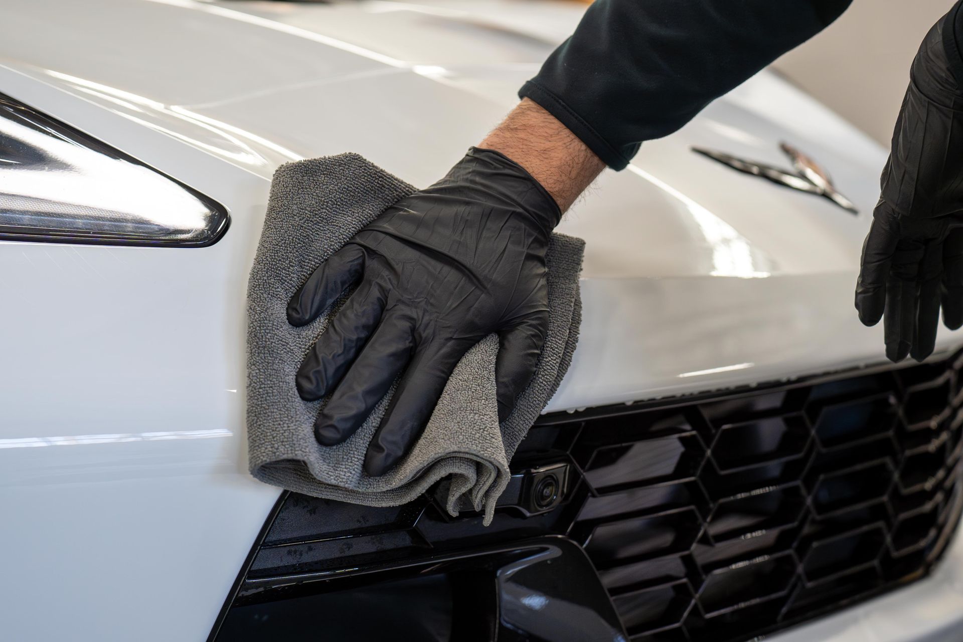 Gloved hands wiping the front grille of a silver car with a gray cloth