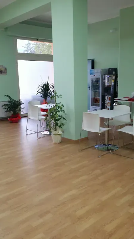 Luminosa stanza moderna con pareti verdi, pavimento in legno, piante in vaso e un tavolo e sedie bianchi accanto a un distributore automatico.