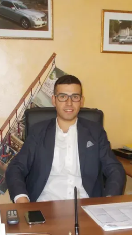 Un uomo in abito blu scuro e occhiali è seduto a una scrivania in un ufficio, con foto incorniciate appese al muro.