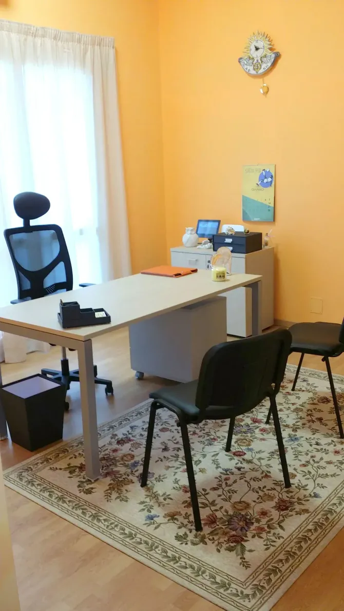Piccolo ufficio con scrivania bianca, sedie nere, monitor e tappeto a motivi geometrici vicino a una finestra.