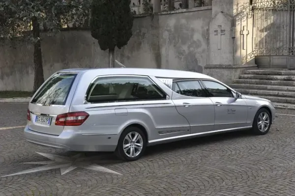 Carro funebre argentato parcheggiato su una strada lastricata di ciottoli accanto a gradini di pietra e un muro