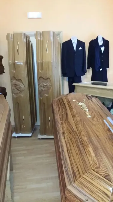 Showroom con bare in legno, abiti blu scuro esposti sulla parete e una bara in legno lucido in primo piano.