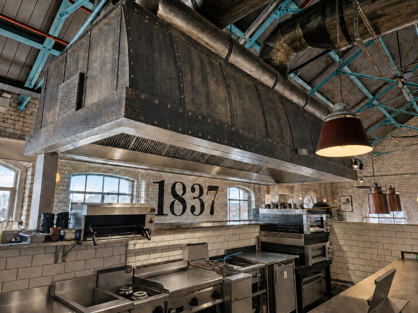 Industrial kitchen with stainless steel counters, brick walls, and large “1837” sign under a high ceiling