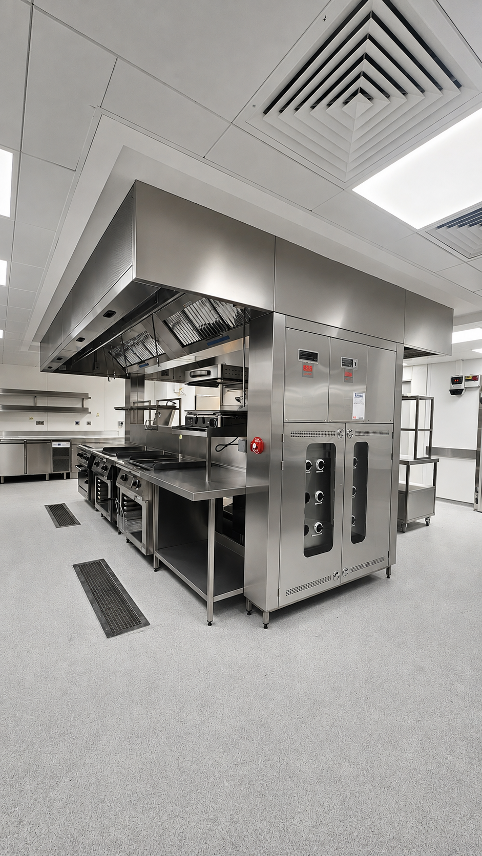 Large stainless-steel industrial kitchen machine in a clean commercial kitchen, with vents and tiled floor.
