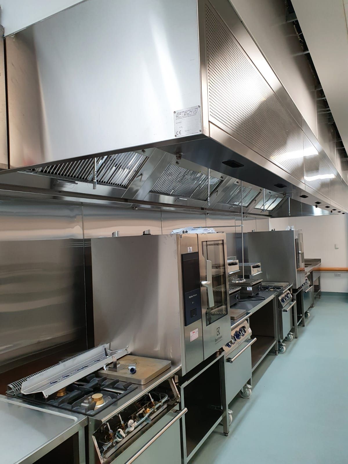 Commercial kitchen with stainless steel cooking equipment and large ventilation hoods along a prep line