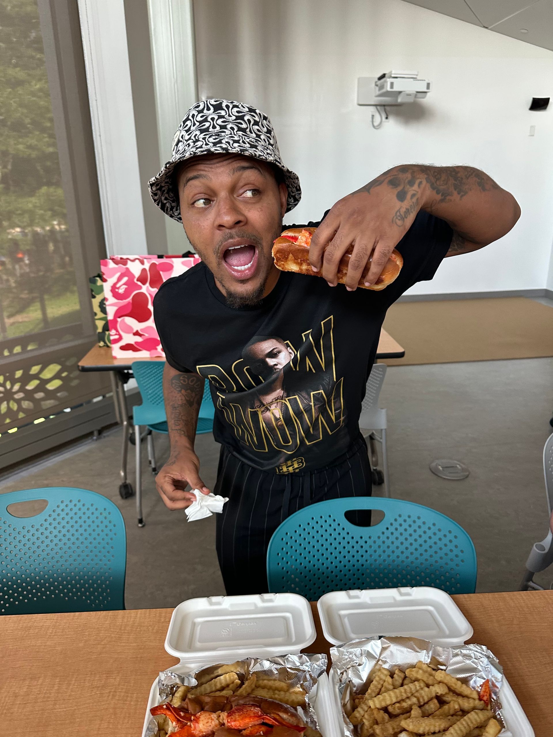 Person at outdoor table eating from takeout containers, wearing a hat and black shirt.