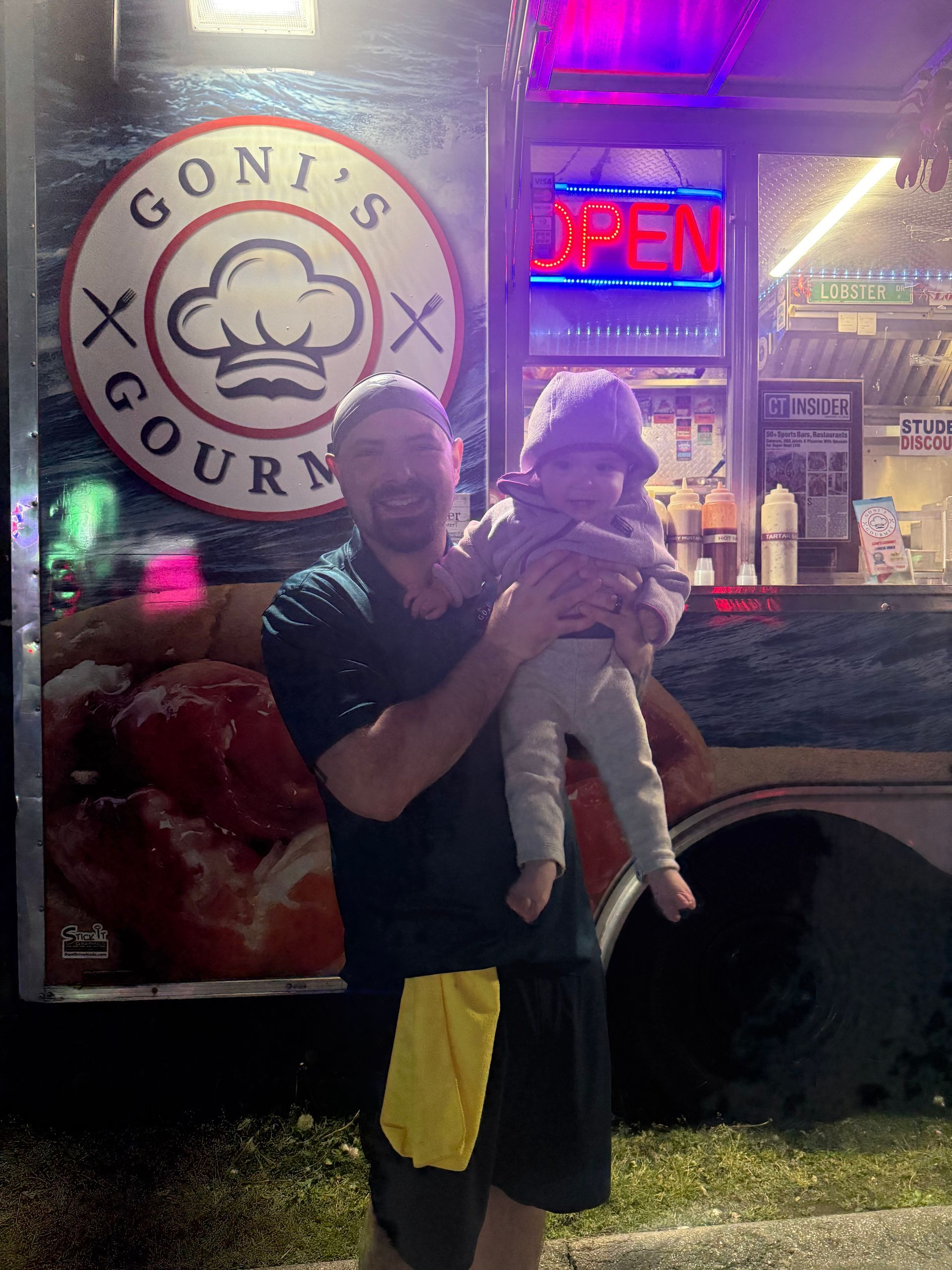 Man holding a baby outside Goni’s Gourmet at night, with neon OPEN sign and colorful lights behind them