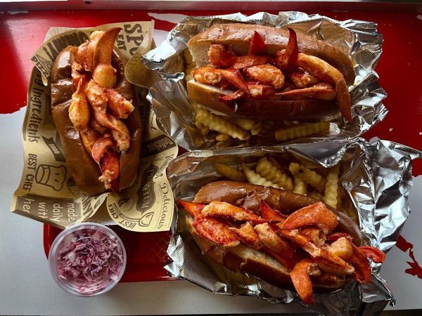 Three foil-lined trays of loaded fries with red sauce and a cup of purple dipping sauce on a red table