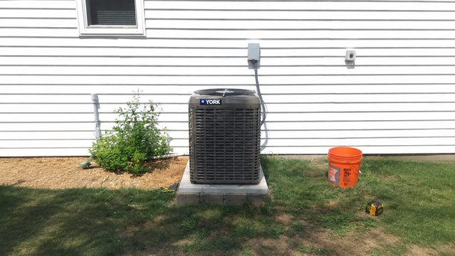Outdoor HVAC unit beside a house with a window, shrub, orange bucket, and lawn.