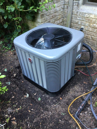 Outdoor air conditioner unit beside a brick wall, with hoses and cables on the ground.