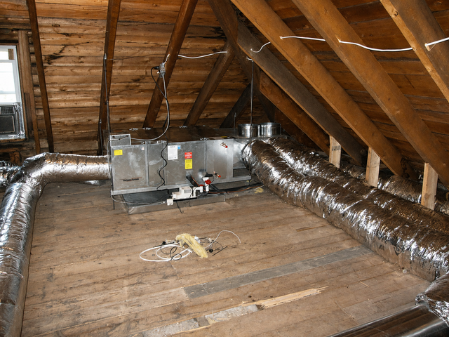 Attic with exposed wooden rafters and silver ductwork, showing an HVAC unit on the floor