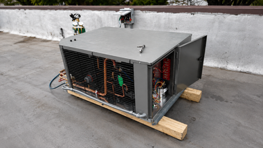 Large metal equipment crate on wooden skids outdoors, with a mesh side panel and cables visible.