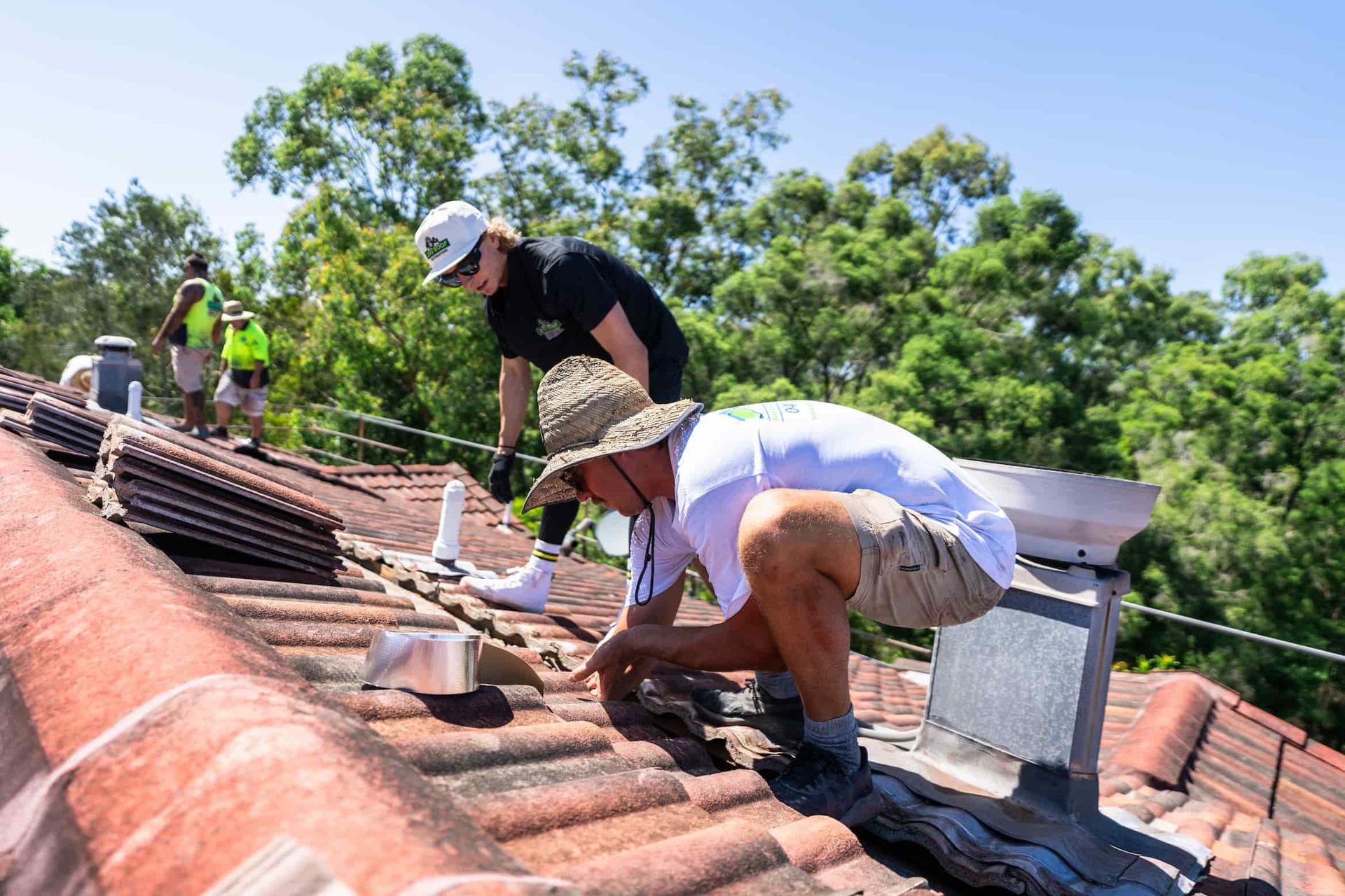Workers repairing a tiled roof on a sunny day, with trees and blue sky in the background