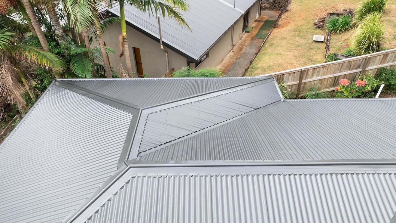 An aerial, high-angle view of a grey corrugated metal roof with various angular sections, next to a house and backyard.