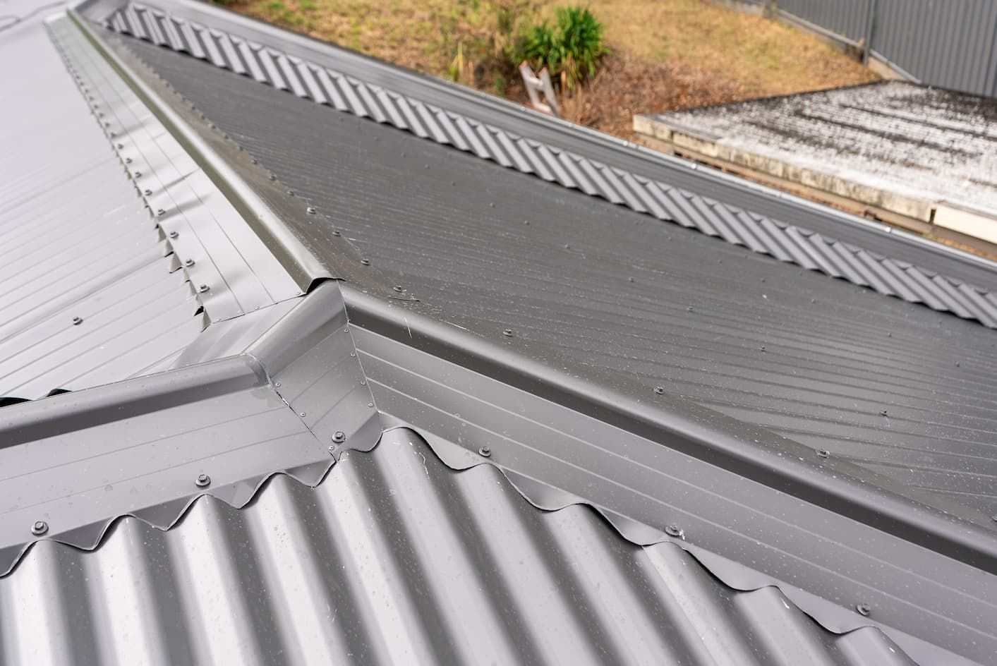 A close-up view of a metal roof with a valley gutter separating a light grey corrugated section from a dark grey surface.