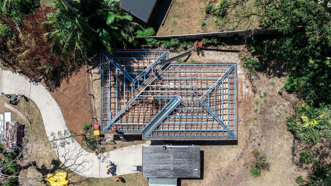An aerial view of a metal roof framing structure being built on a construction site surrounded by trees and a walkway.