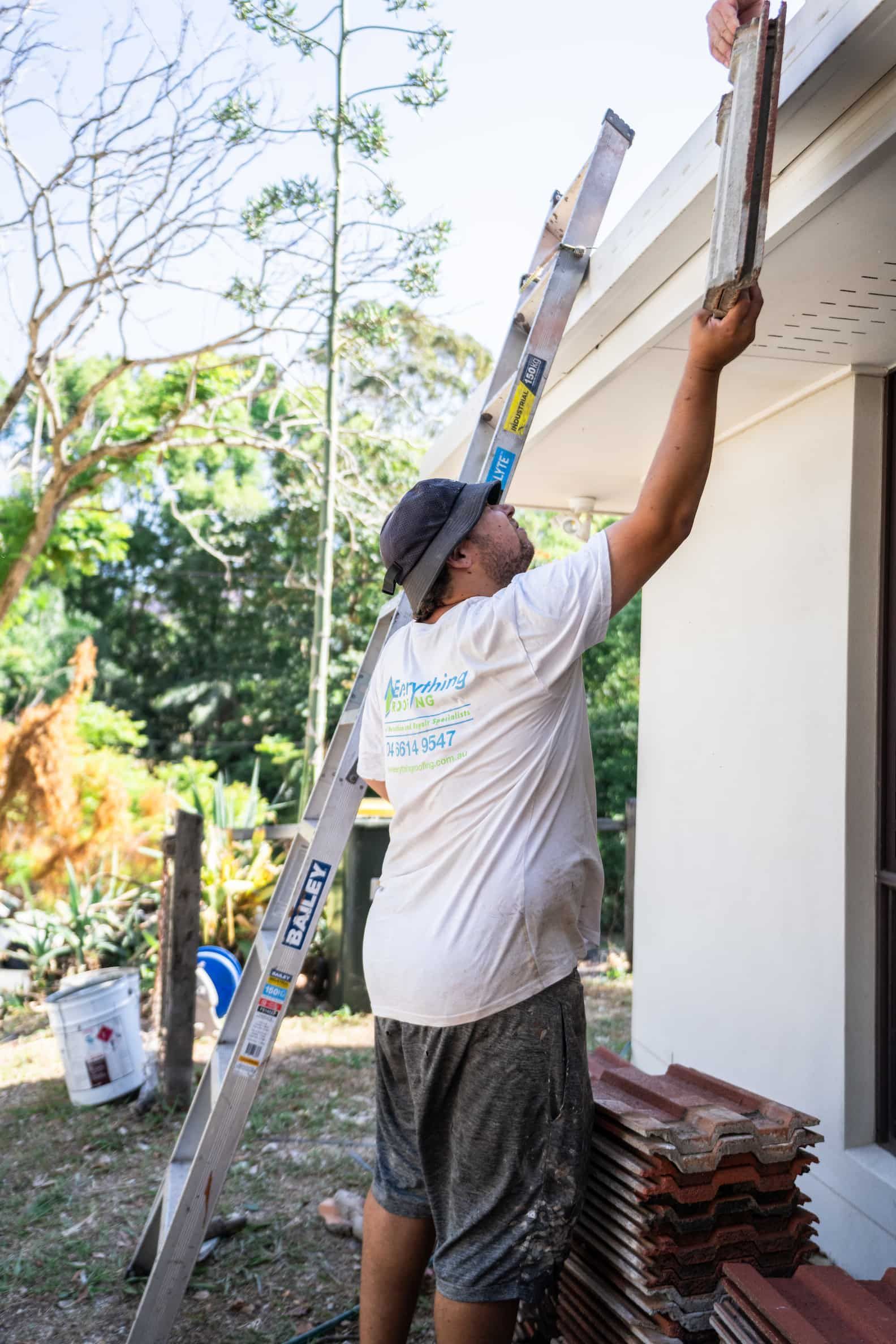 A person in a hat and white shirt stands on a ladder, installing a brown tile on the roof edge of a house.