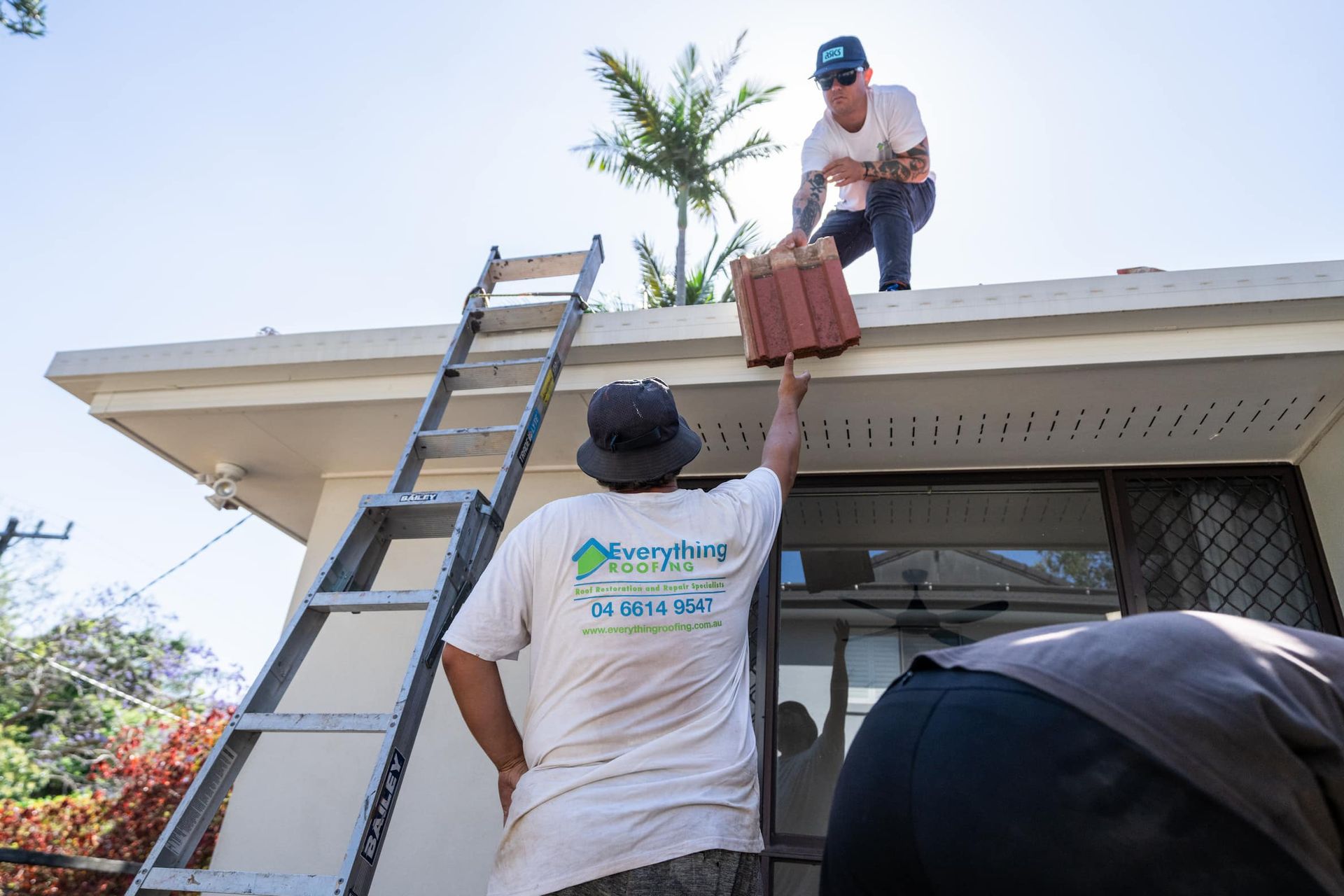 A worker on a roof hands a stack of red tiles to a colleague standing on the ground near a tall ladder.