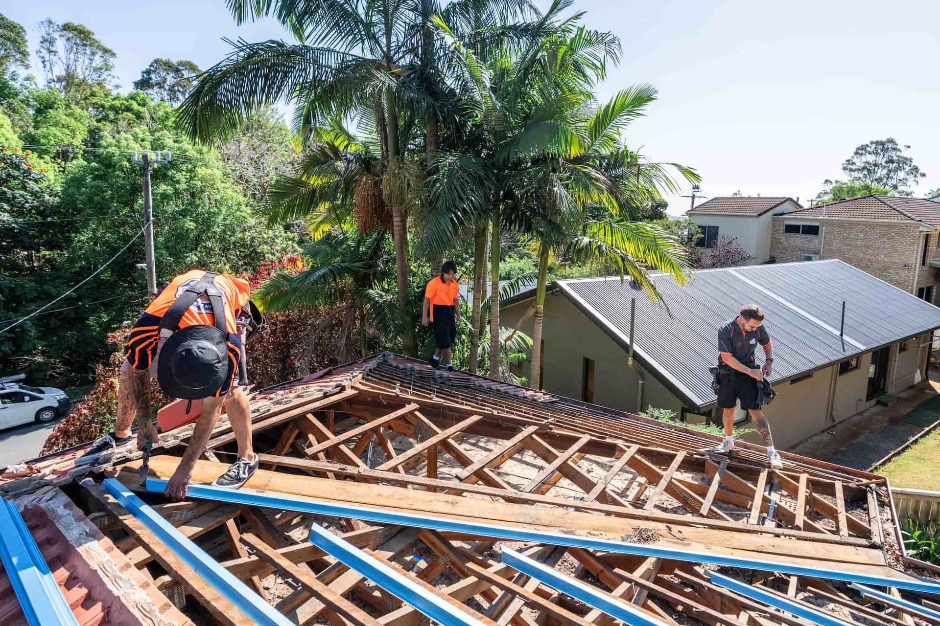 Three people in work clothes repair a roof with an exposed wooden frame, working with blue metal beams on a sunny day.