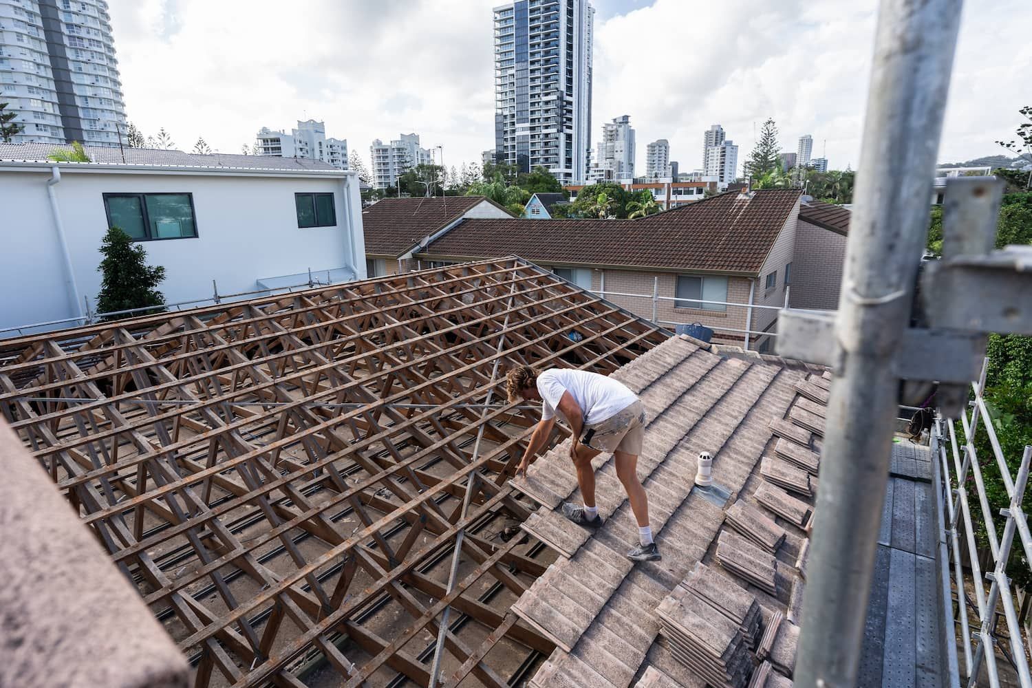 A person works on a roof, installing tiles over wooden trusses while overlooking a city neighborhood with tall buildings.